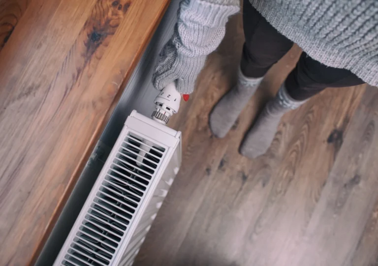 Person wearing gloves and wool socks adjusts the temperature dial on a white radiator in a room with wooden flooring.