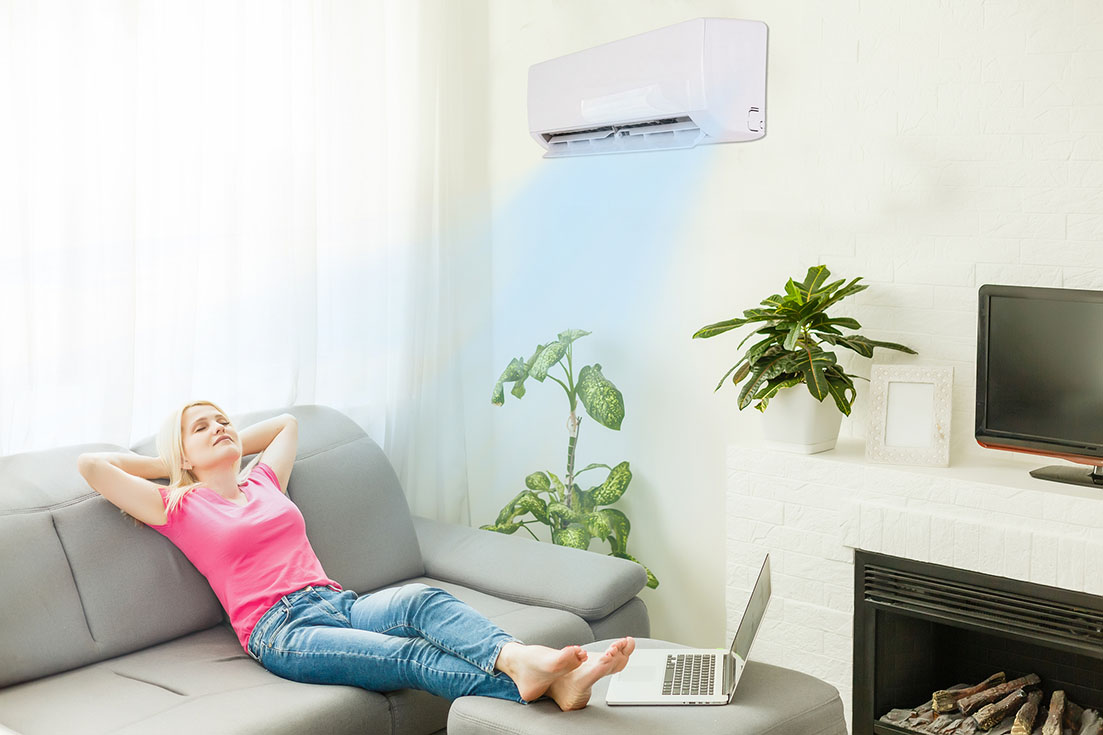 Young happy woman switching on air conditioner sitting on couch at convenient cozy home, lady relaxing on sofa in living room holding remote climate control to cooler system set comfort temperature