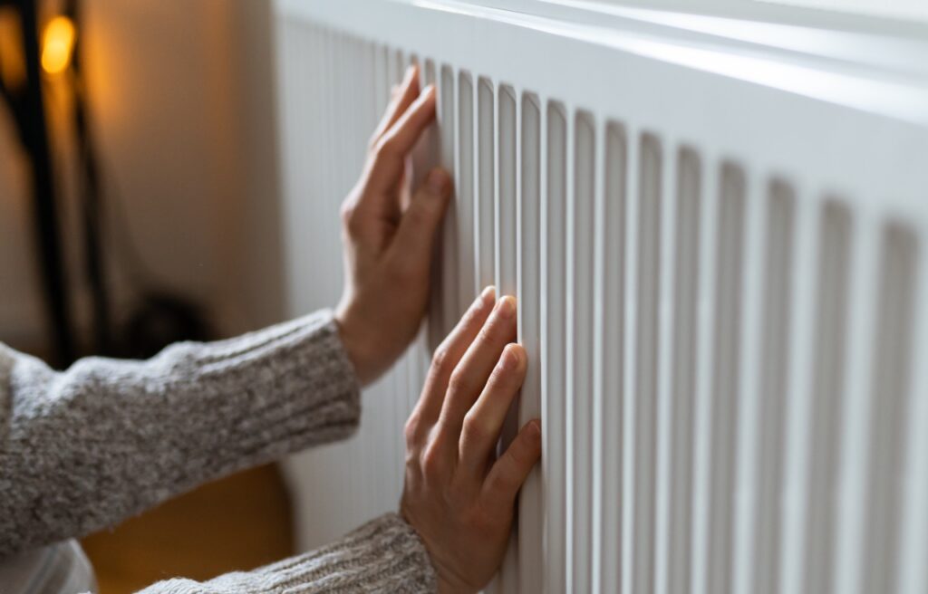 A person in a gray sweater places both hands on a white radiator, likely to feel warmth.